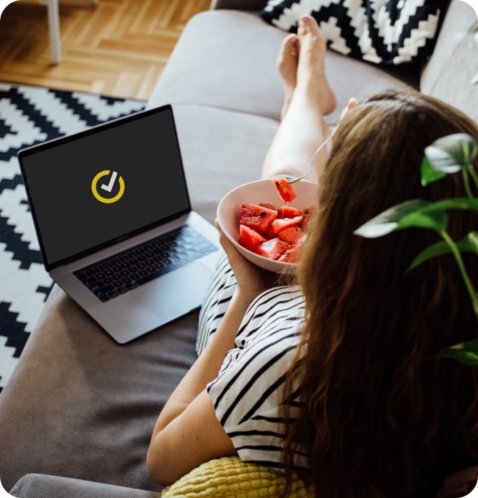 Nő görögdinnyét eszik a kanapén, miközben a laptopon elindul a Norton 360 AntiVirus Plus